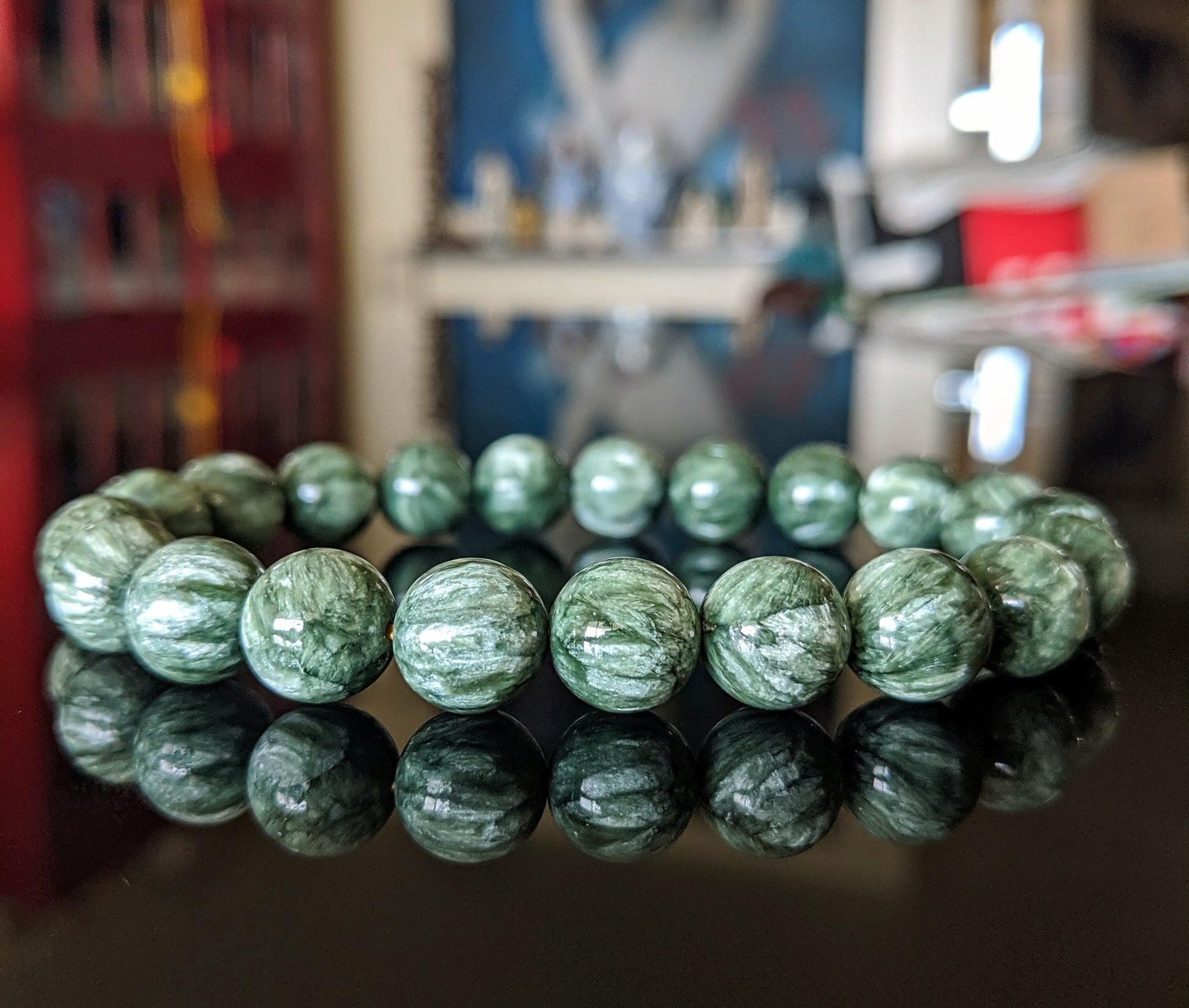 This is the main picture for the  Seraphinite bead bracelet 10mm.  It is 3A+ , the highest grade available on the market.  The bracelet is displayed on a black flat surface.  it is a dark green with marblized bands of white.  It shimmers under the light.  