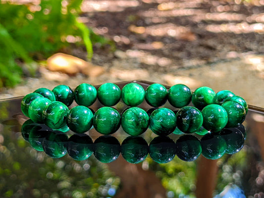 This is the main picture for the Green Tiger Eye bead bracelet.  It is displayed on a flat black surface with nature on the background.  The quality of this Green Tiger Eye is superb.  It has a polished finished and the green color is vibrant. 