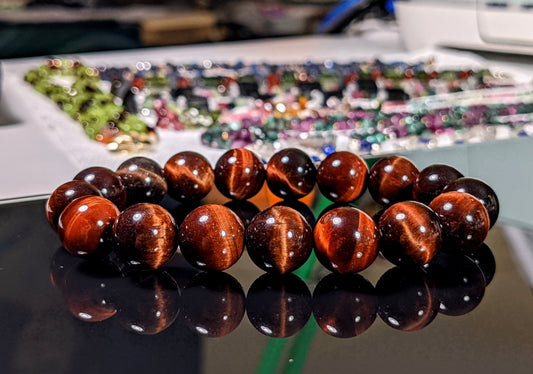 On this image, the Red Tiger eye bead bracelet is displayed on a black flat surface.  The Red Tiger Eye is 3A quality; excellent grade.  The beads are 12 mm size, and they are strung on durable stretch cord.  RayonBracelet.com