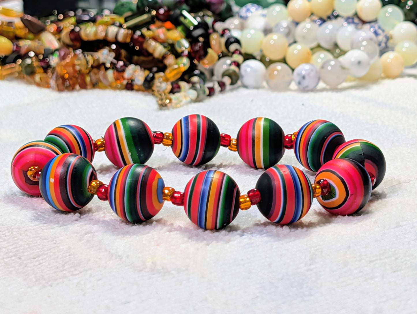 A beaded stretch bracelet made of round striped soft clay beads in vibrant colors including pink, green, blue, yellow, orange, and black, separated by small red and yellow spacer beads, displayed on a white textured surface with other jewelry blurred in the background.